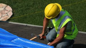 Ouvrier en casque jaune posant une bâche bleue de protection sur un toit endommagé